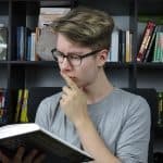 Young man with glasses contemplating a book in a library setting.