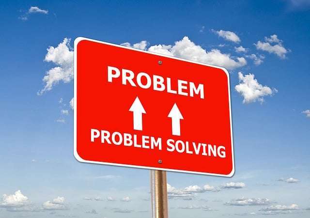 A bright red rectangular sign against a blue sky with scattered white clouds. The sign features the word "PROBLEM" at the top and "PROBLEM SOLVING" at the bottom in bold white capital letters. Two white arrows point upward from the bottom phrase toward the top word, symbolizing a direct approach to overcoming challenges.