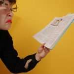 Woman with glasses making a comical, confused face while peering closely at an open book.