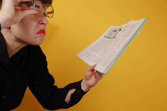 Woman with glasses making a comical, confused face while peering closely at an open book.