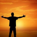 Silhouette of a person standing on the beach with open arms during a warm sunset, representing simple ways to find joy and happiness in everyday life.